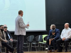 Man on stage talking to another man in front of a large screen